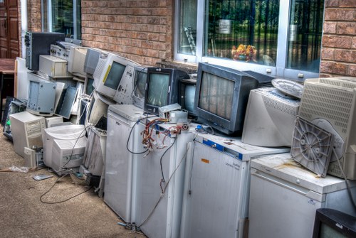 A row of discarded old computer monitors and desktop units arranged on the pavement outside a residential building with brick walls and a window. The electronic equipment varies in size, with some monitors featuring thick bezels and others being flat-screen types, while the desktop towers show a mix of beige, grey, and black finishes. Wires and cables are loosely connected or hanging from some of the units, and a few are stacked or leaning against each other. The setting appears to be the driveway or front area of a house in Newham, with a partial view of a window reflecting greenery. The collection of electronic waste is in need of removal and recycling, an activity serviced by local rubbish removal specialists like Newham House Clearance. The environment is illuminated by natural daylight, casting soft shadows on the ground and creating a neutral, professional atmosphere suitable for a rubbish disposal context.