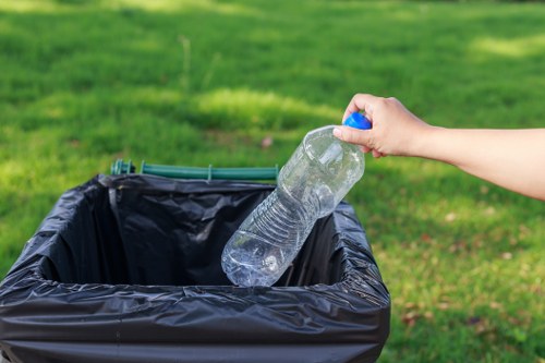 A person's hand holding a clear plastic water bottle with a blue cap, about to dispose of it into a black plastic rubbish bin lined with a black trash bag, set outdoors on a paved area adjacent to a well-maintained grassy lawn. The background features lush green grass and a blurred natural environment, suggesting an outdoor location typical of residential or commercial rubbish collection sites in Newham. The scene reflects general rubbish disposal and waste management practices, with focus on recycling or waste separation efforts undertaken by local rubbish removal services such as Newham House Clearance.
