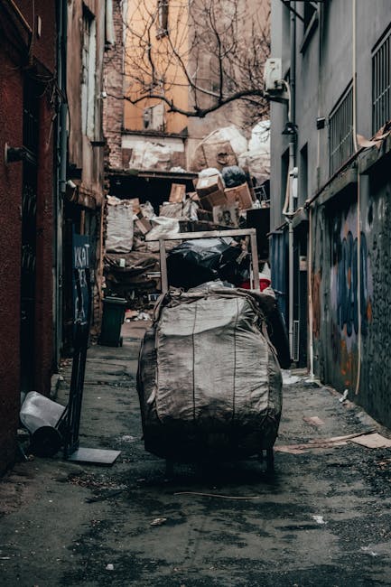 An outdoor scene shows three individuals amidst a large accumulation of mixed waste and rubbish on uneven ground. The focus is on a white delivery or storage container with the identification number '