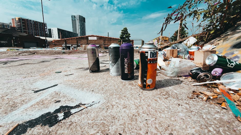 A narrow alleyway in an urban area, featuring uneven, muddy ground with patches of dampness and scattered debris. The left side is lined with weathered brick walls covered in various colourful graffit