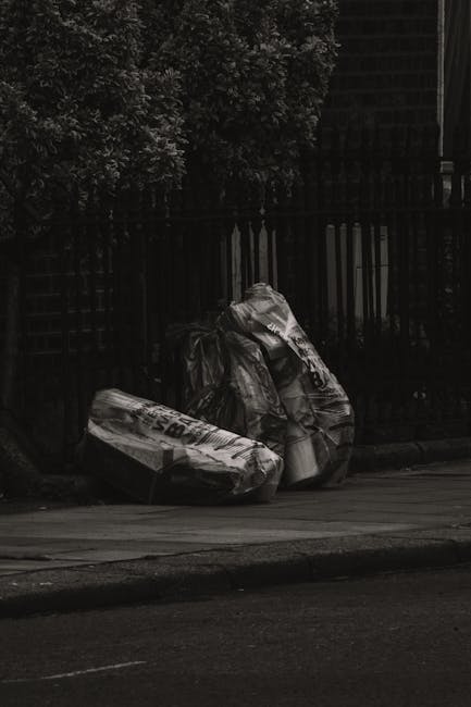 A collection of overflowing waste and rubbish bags positioned on a paved sidewalk in front of a building with commercial units, including a fish bar. The rubbish includes black plastic bin bags, cardb